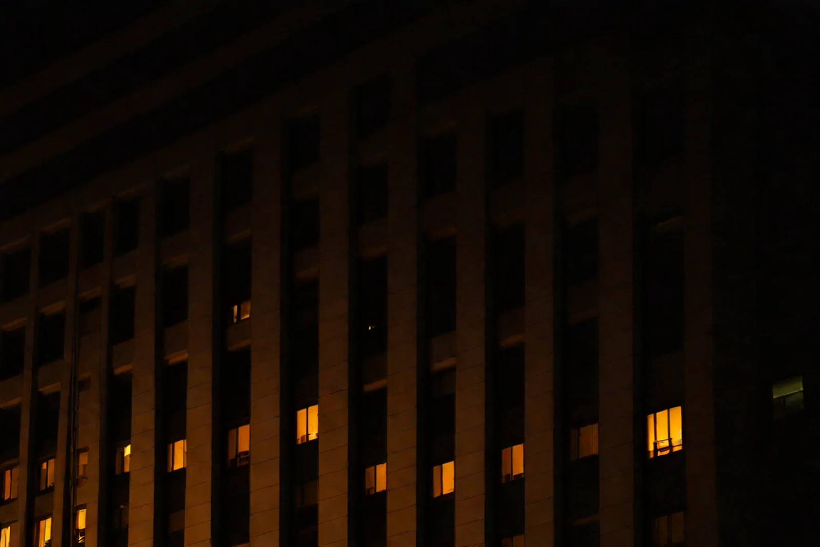 Fachada de un edificio de noche: casi todas las ventanas apagadas y unas pocas encendidas en ámbar.